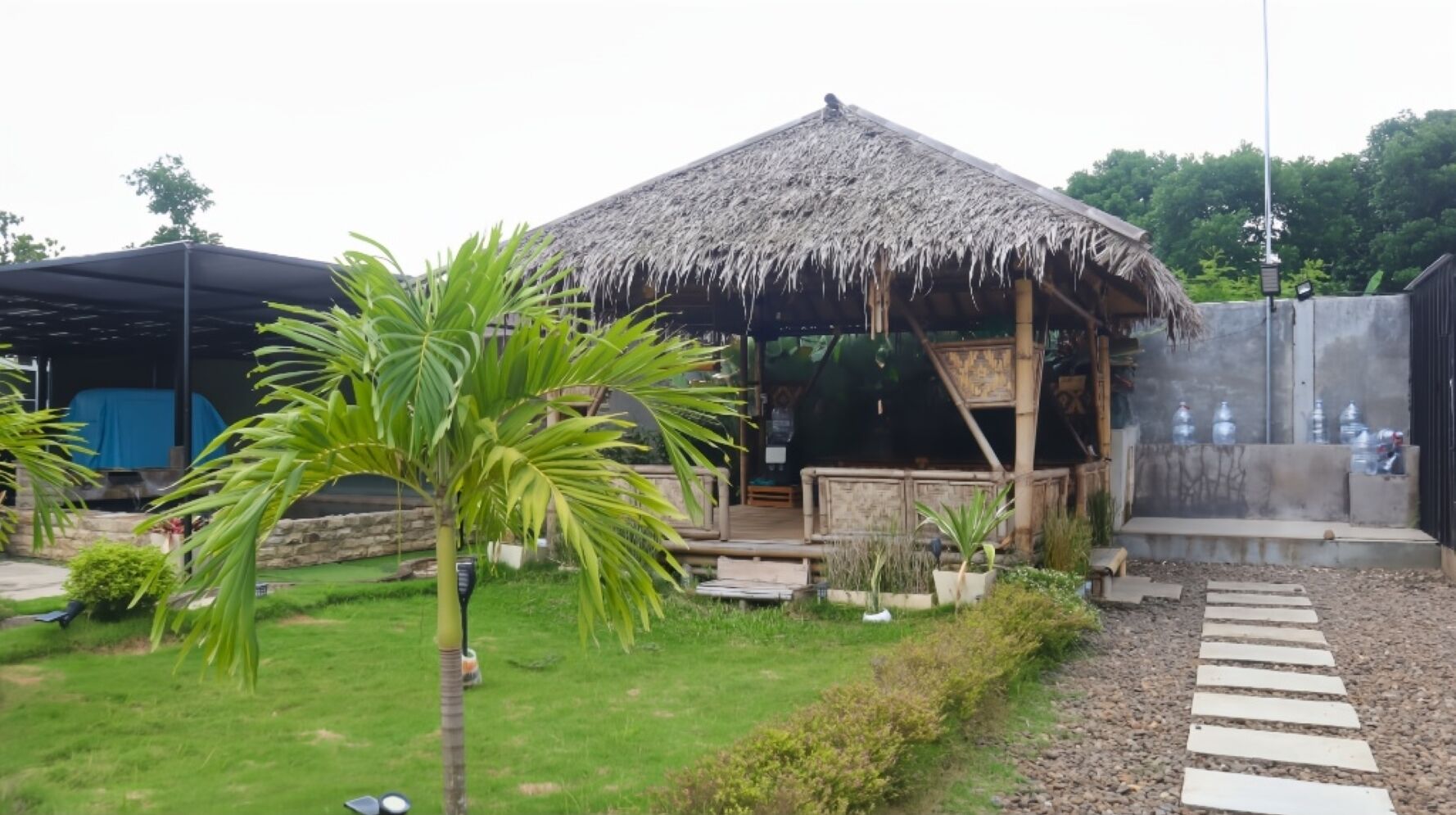 Una estructura con techo de paja y terraza de madera, rodeada de follaje y un camino de grava.