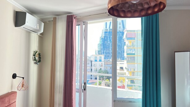 A room with a large window, a white air conditioning unit, a pink sofa, a wall-mounted light fixture, and a white refrigerator. - SUNLIT ERISI’s HOUSE - City center (Tiranë)