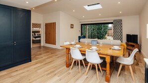 A spacious dining area with a wooden table and white chairs, a dark door, and a view of a garden through a sliding door. - Cart Shed 2 - From 140 Per Night (Oxford)