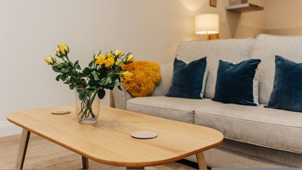 A living room with a wooden coffee table, a vase with yellow flowers, a beige sofa with blue cushions, and a yellow throw blanket. - Cart Shed 2 - From 140 Per Night (Oxford)