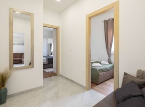 A modern hotel room with a large mirror, a bed with a green bedspread, and a view of a cityscape through the window. - Contemporary Apartment With Terrace and Spacious Garden (Kastela)
