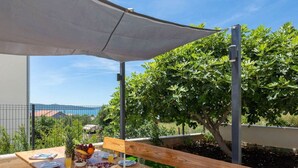 A wooden table set with plates, glasses, and a basket of croissants under a white canopy, overlooking a garden with trees and a view of the sea.