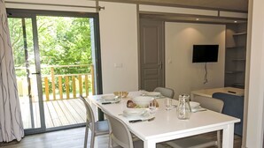 Ein Essbereich mit einem weißen Tisch, gedeckt für vier Personen, einer Holzterrasse davor und einem an der Wand montierten Fernseher.
