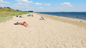 Ein sandiger Strand mit badenden und sonnenden Personen, ein klarer, blauer Himmel und sanfte Hügel in der Ferne.