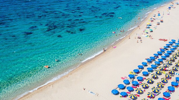 Una playa con agua turquesa cristalina, una orilla arenosa y numerosos paraguas azules y amarillos.
