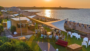 Un espacio al aire libre junto a la costa con mesas y sillas, una terraza de madera y vistas al mar y al atardecer.