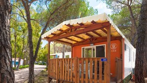 Una cabaña pequeña de una sola planta con terraza de madera y porche cubierto, rodeada de árboles.