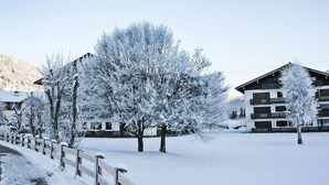 A snow-covered landscape with a wooden fence, a tree, and a building with multiple balconies. - Dachstein Top 12 by Interhome (Flachau)