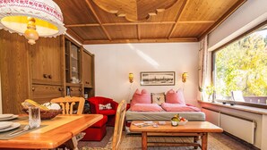 A cozy room with a wooden ceiling, a dining table with chairs, a sofa with red and white checkered pillows, and a window with a view of trees. - Dachstein Top 12 by Interhome (Flachau)