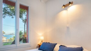 A bedroom with a large bed, two pillows, a nightstand with a lamp, and a window with a view of the sea. - Le Provençal by Interhome (Le Pradet)