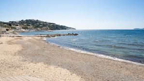 A beach with pebbles, a wooden walkway, and a distant hill with buildings. - Le Provençal by Interhome (Le Pradet)