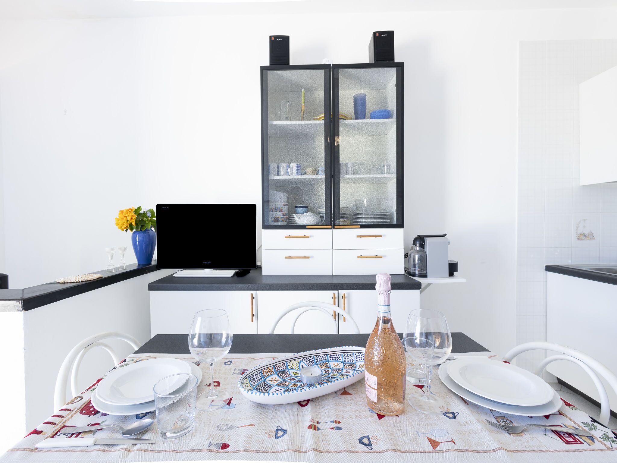 A modern kitchen with white cabinetry, a black countertop, and a dining table set with plates, glasses, and a decorative vase.