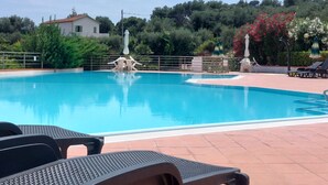 Una piscina con sillones de descanso y sombrillas, rodeada de follaje y un edificio al fondo.