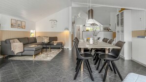 A modern living room with a grey sofa, a dining area with a white table and black chairs, and a kitchen in the background.