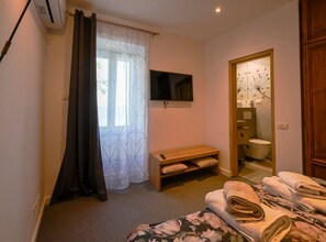 Un dormitorio con cama, un banco, televisor y baño con inodoro y cortina de ducha.