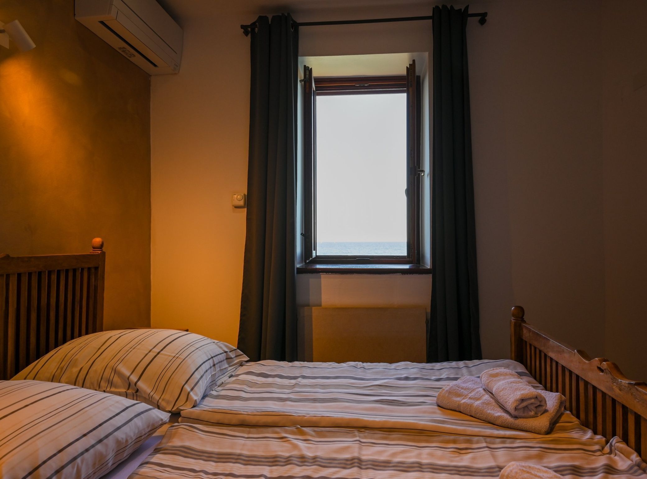 Un lit simple avec une literie à rayures et une serviette enroulée, une fenêtre avec des rideaux et un appareil de climatisation fixé au mur.