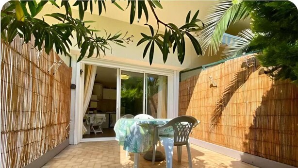 A covered patio with a table and chairs, a sliding glass door leading to a kitchen, and a bamboo fence. - Cocon au Pradet - Beach, terrace & absolute calm (Le Pradet)