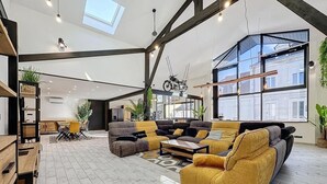 Ein geräumiger Wohnbereich mit einer großen Couch, einem Couchtisch und einem Esstisch. Pflanzen, ein Fahrrad und ein Ausblick ins Freie durch große Fenster sind zu sehen.