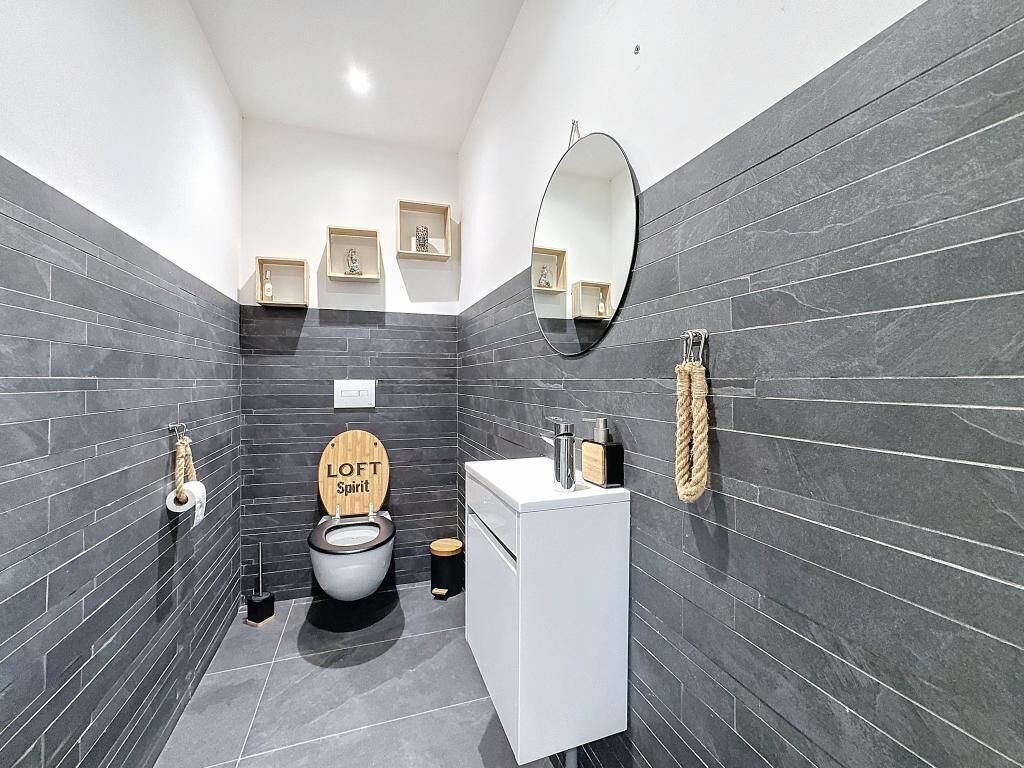 Une salle de bain moderne avec des murs recouverts de carreaux foncés, un lavabo blanc, des toilettes avec un abattant en bois et un miroir.