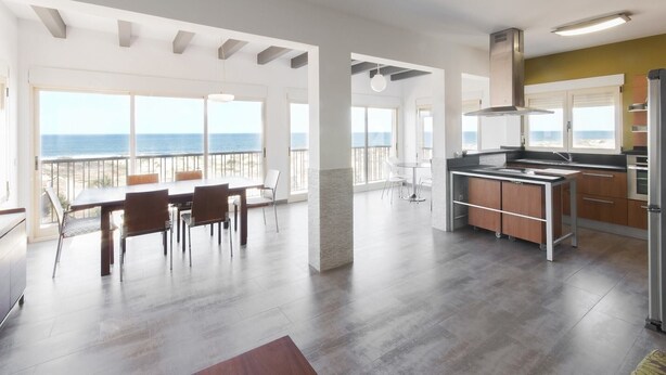 Et rummeligt køkken med havudsigt, et spiseområde med et stort bord og stole, samt et moderne design med synlige bjælker og store vinduer.