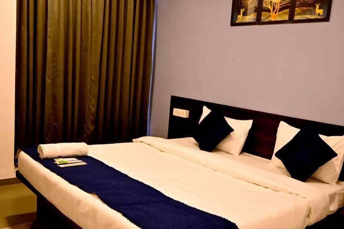 A neatly made bed with a dark headboard, a blue throw blanket, and white bedding. There is a painting on the wall above the bed, and a window with dark curtains.