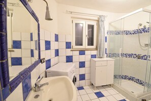 Un baño con lavamanos blanco, lavadora, espejo y ducha con mampara de vidrio.