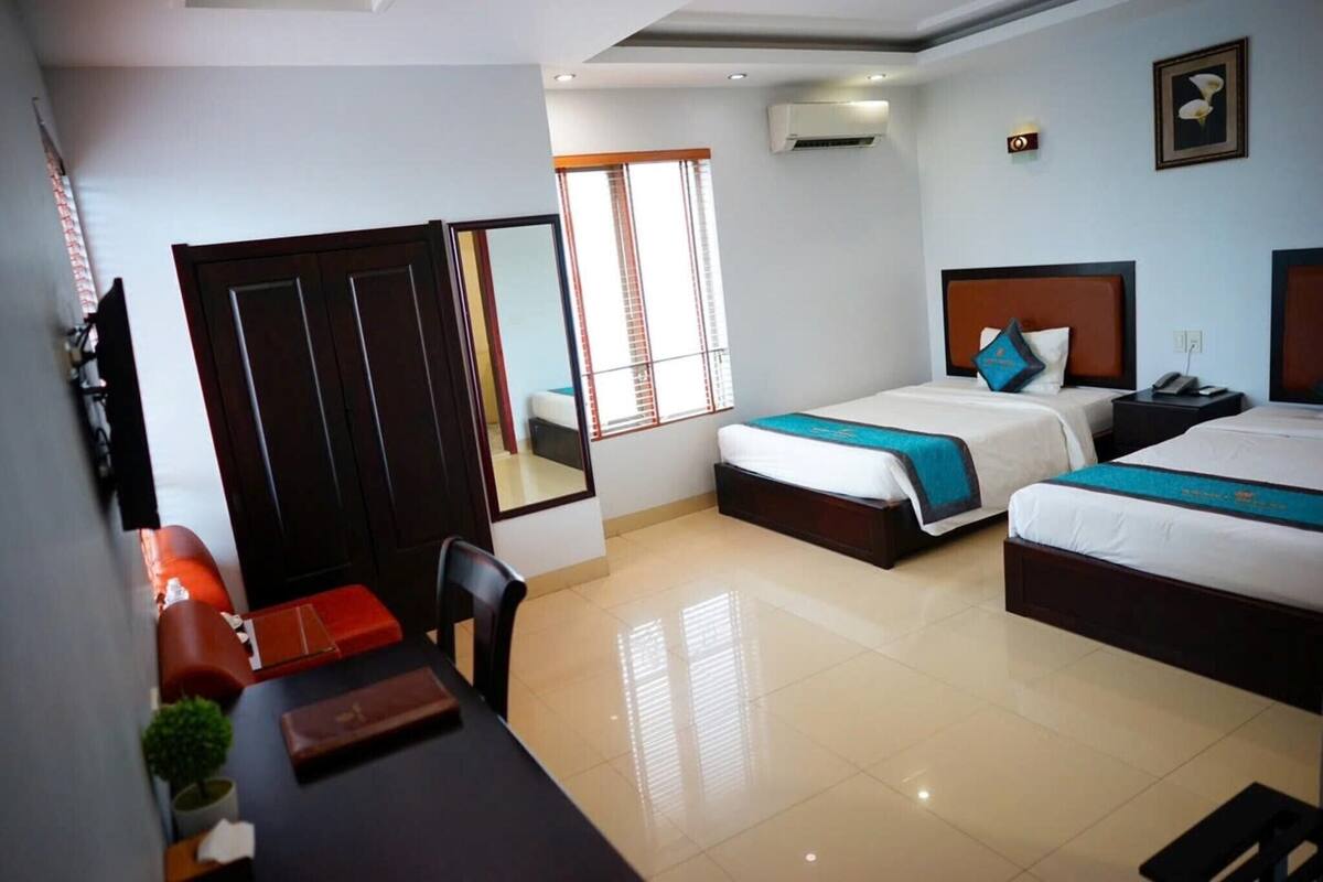 A hotel room with two beds, a desk, a chair, a wardrobe, and a window with blinds.