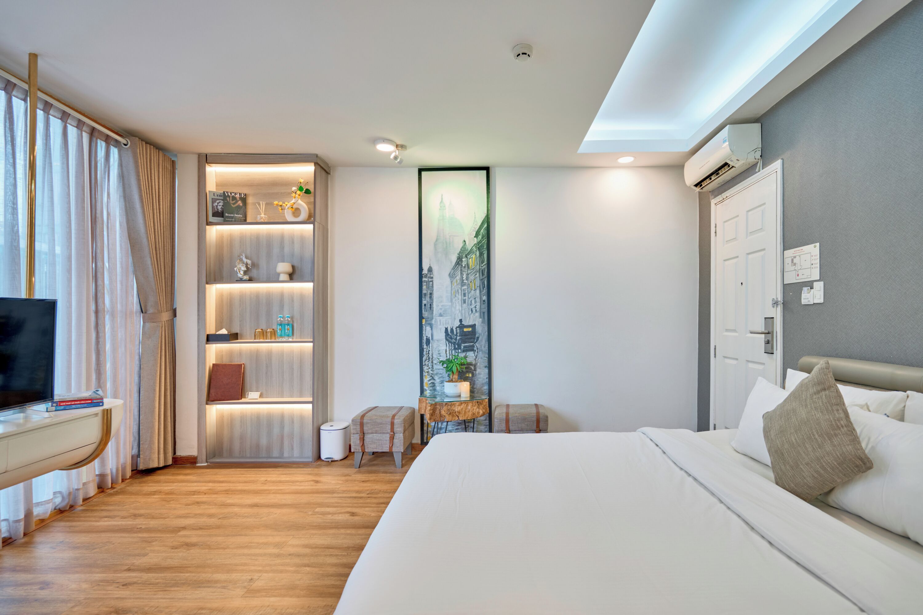 A hotel room with a bed, a TV, a desk, a bookshelf, and a large window with curtains.