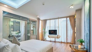 A modern hotel room with a large bed, a TV, a glass shower enclosure, and a view of the city.