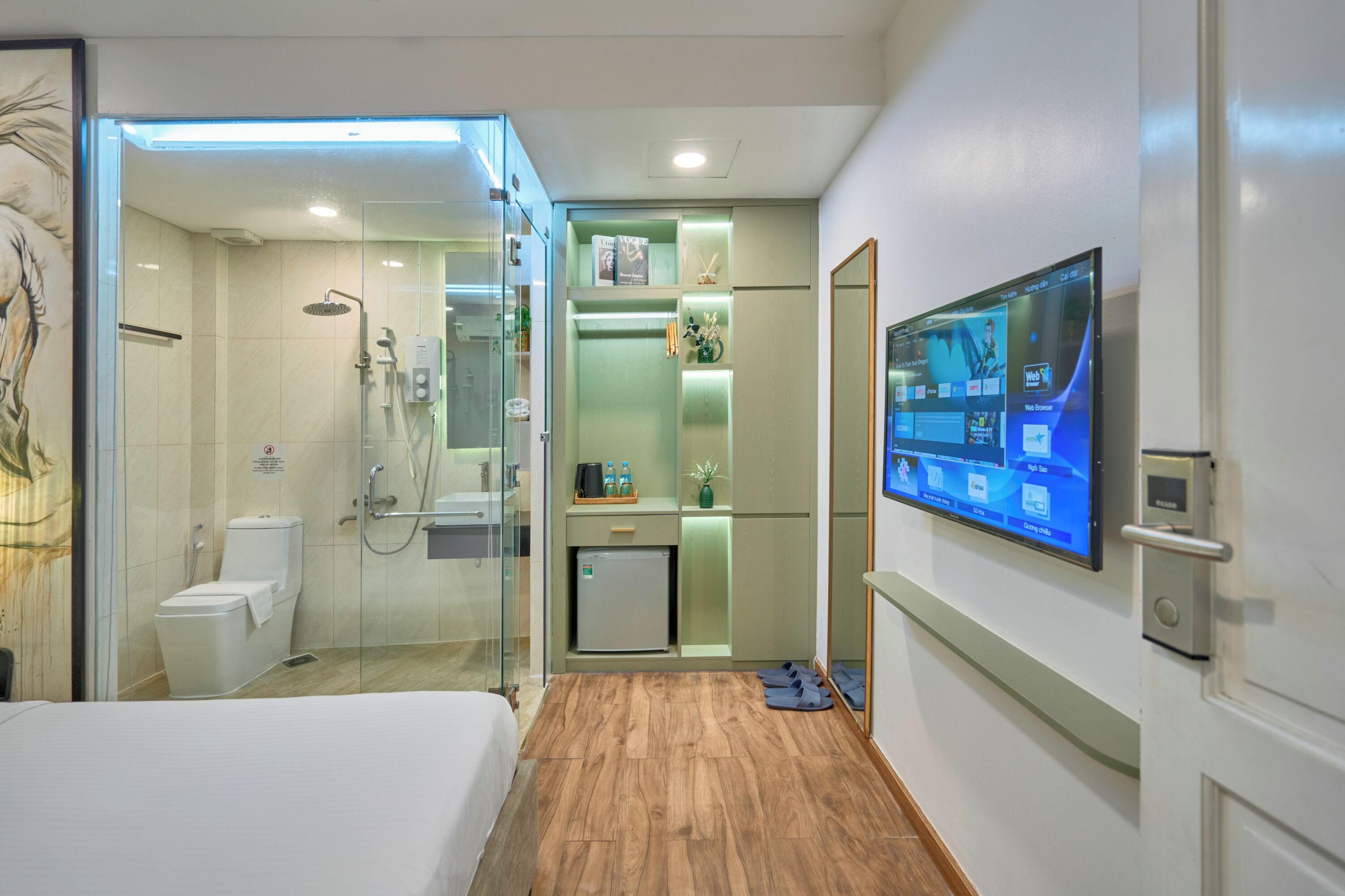 Una habitación de hotel moderna con cabina de ducha de vidrio, cama, refrigerador pequeño, televisor montado en la pared y piso de madera.