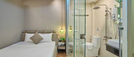Une chambre d’hôtel moderne équipée d’un lit, d’une table de chevet, d’une lampe et d’une douche séparée par des parois en verre.