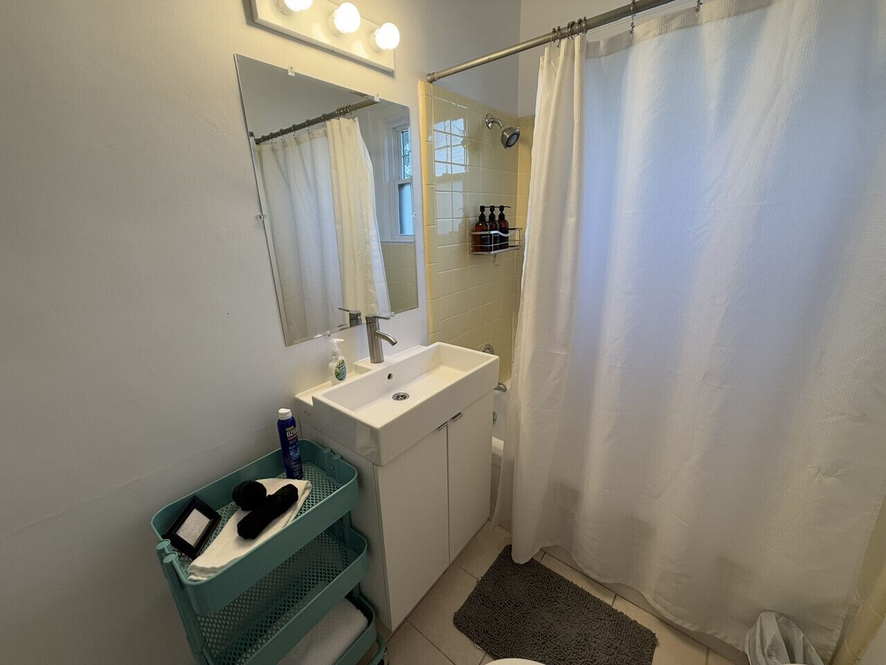 A bathroom with a white sink, mirror, and shower curtain. There is a green basket and a black object on the floor.