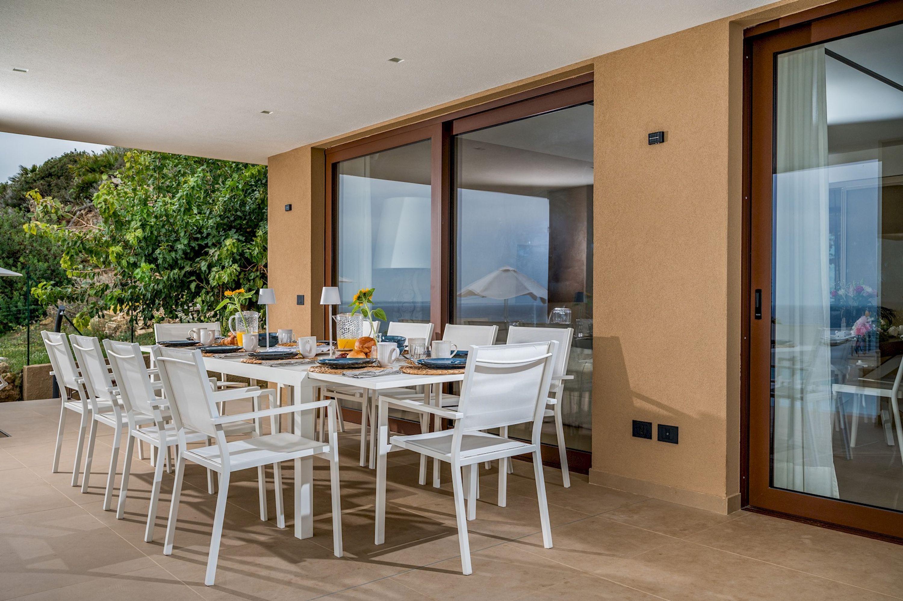 A modern outdoor dining area with a white table and chairs, a glass sliding door, and a view of the garden.