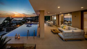 A modern infinity pool with a sunset view, a cozy seating area with a sofa and chairs, and a well-lit interior space.
