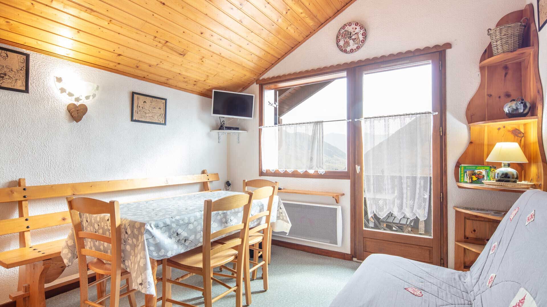 A cozy room with a wooden ceiling, a dining table with chairs, a TV mounted on the wall, and a window with a view of the outdoors.