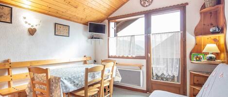 A cozy room with a wooden ceiling, a dining table with chairs, a TV mounted on the wall, and a window with a view of the outdoors.