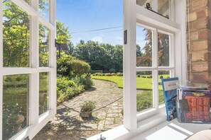 Blick aus einem Fenster auf einen gepflasterten Weg, üppiges Grün und einen klaren, blauen Himmel.