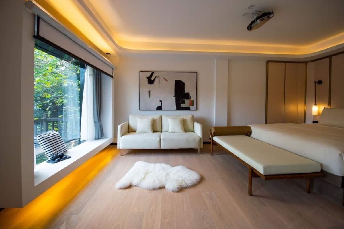 Ein modernes Hotelzimmer mit einem großen Bett, einem Sofa, einer Bank, einem Fellteppich und einem Fenster mit Blick auf Grünflächen.