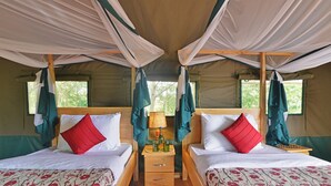 Een tent met twee bedden, elk met een houten frame, wit beddengoed, rode kussens en een klein nachtkastje met een lamp.