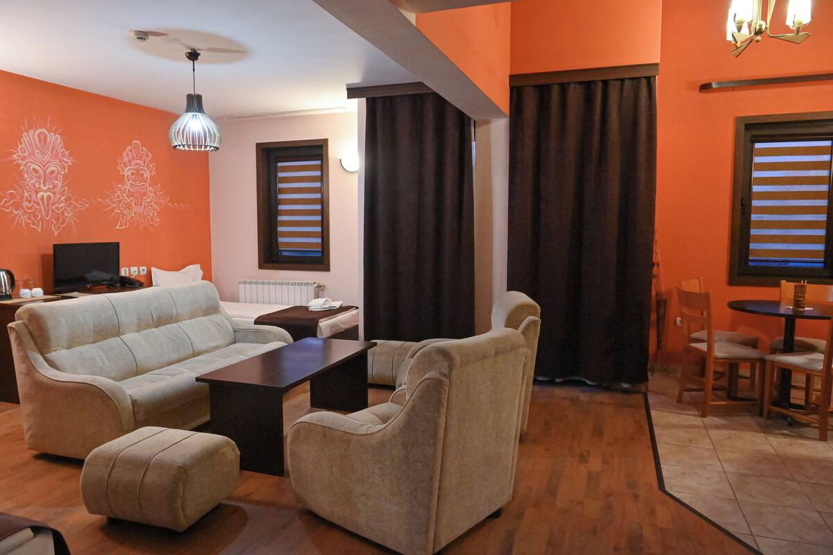 Ein Wohnzimmer mit einer Couch, Sesseln, einem Couchtisch und einem Fernseher. Vorhanden sind Vorhänge, ein Essbereich mit Stühlen und ein Fenster mit Jalousien.