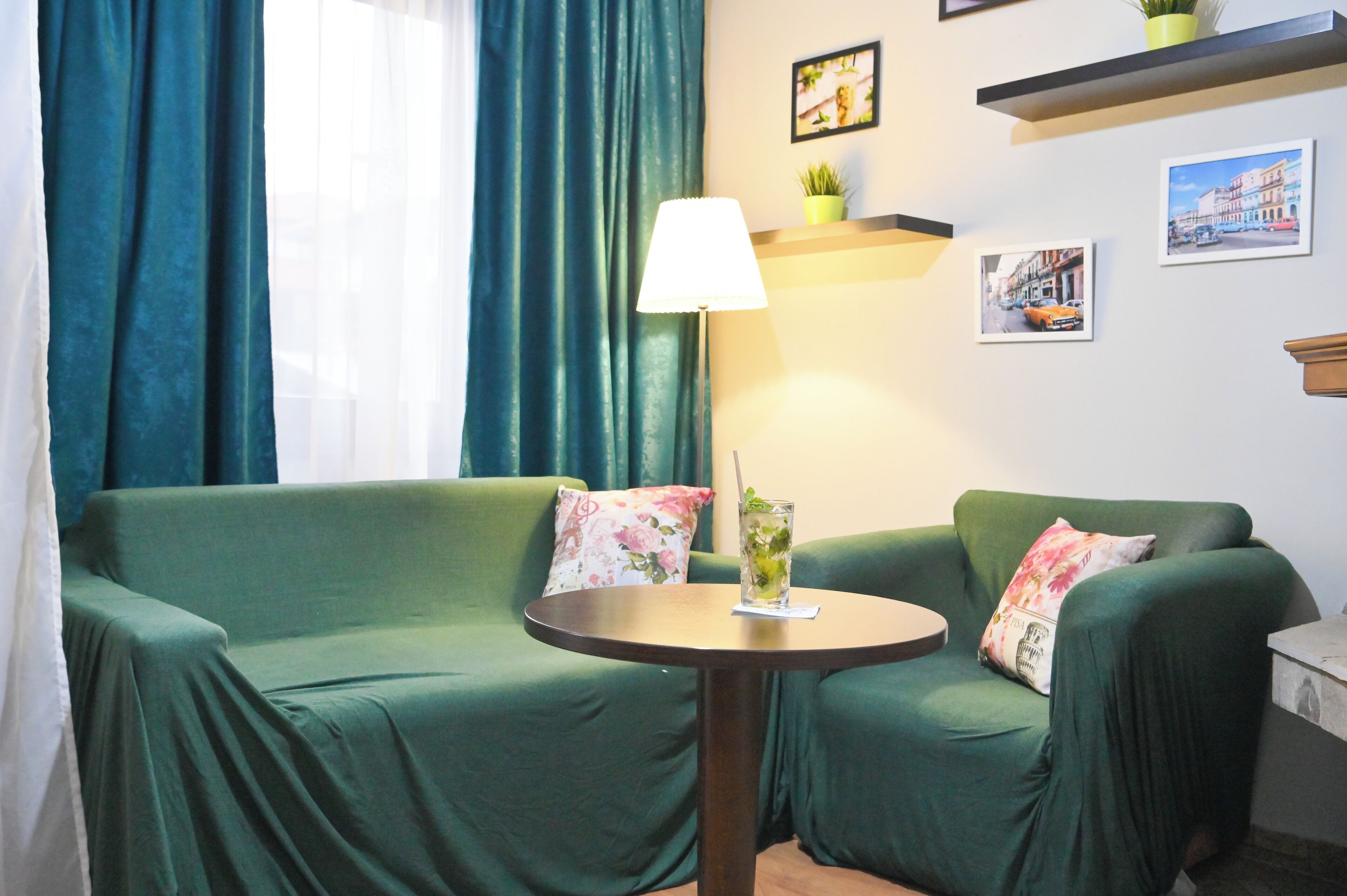 Un canapé vert agrémenté de coussins à motifs floraux, une petite table ronde avec un verre d’eau et une lampe, et un mur orné de tableaux encadrés.