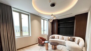 Un salon moderne avec un canapé incurvé, un fauteuil rouge et une table basse. De grandes fenêtres laissent entrer la lumière naturelle et offrent une vue sur les bâtiments environnants.