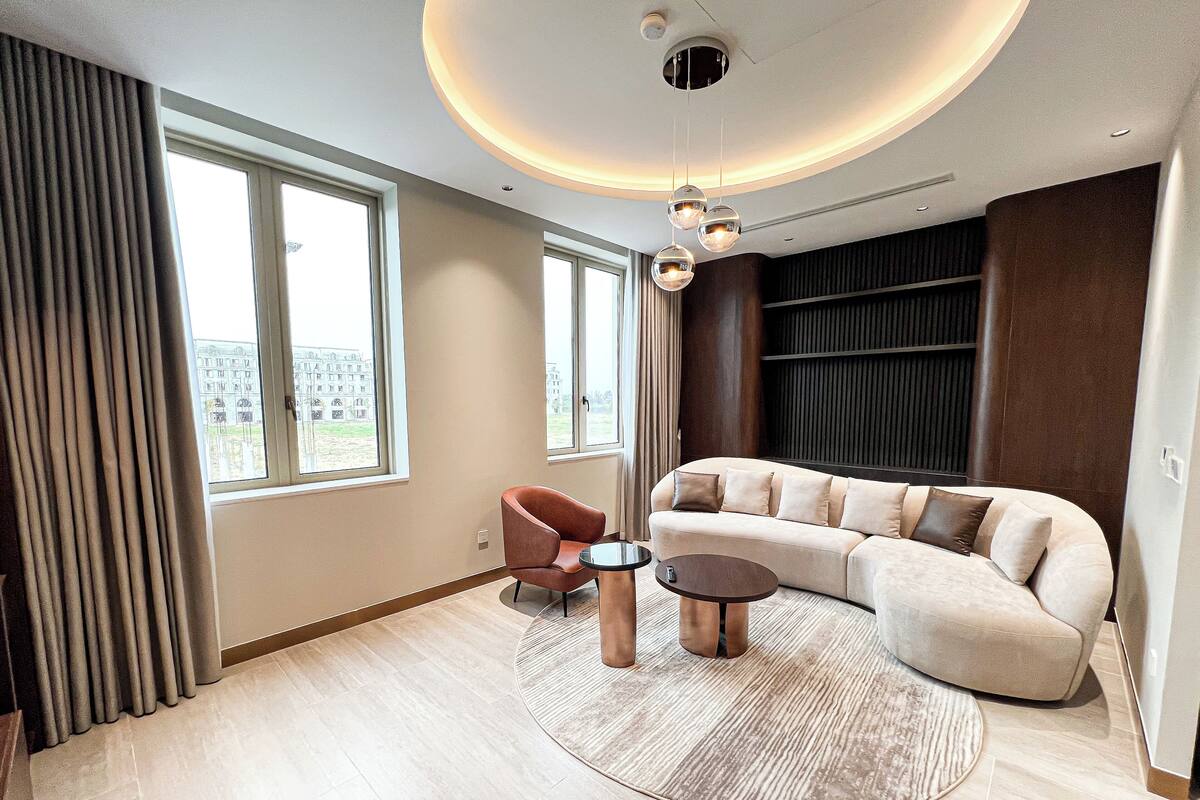 Ein modernes Wohnzimmer mit einer gebogenen Couch, einem roten Sessel und einem Couchtisch. Große Fenster sorgen für natürliches Licht und bieten einen Blick auf Gebäude.