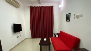 A living room with a red sofa, a small table, a ceiling fan, a flat-screen TV, and a red curtain. - AZAIBA HOTEL APARTMENT (Muscat)