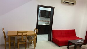 A compact living space with a dining table and chairs, a red sofa, and a small table. A kitchen is visible through an open doorway. - AZAIBA HOTEL APARTMENT (Muscat)