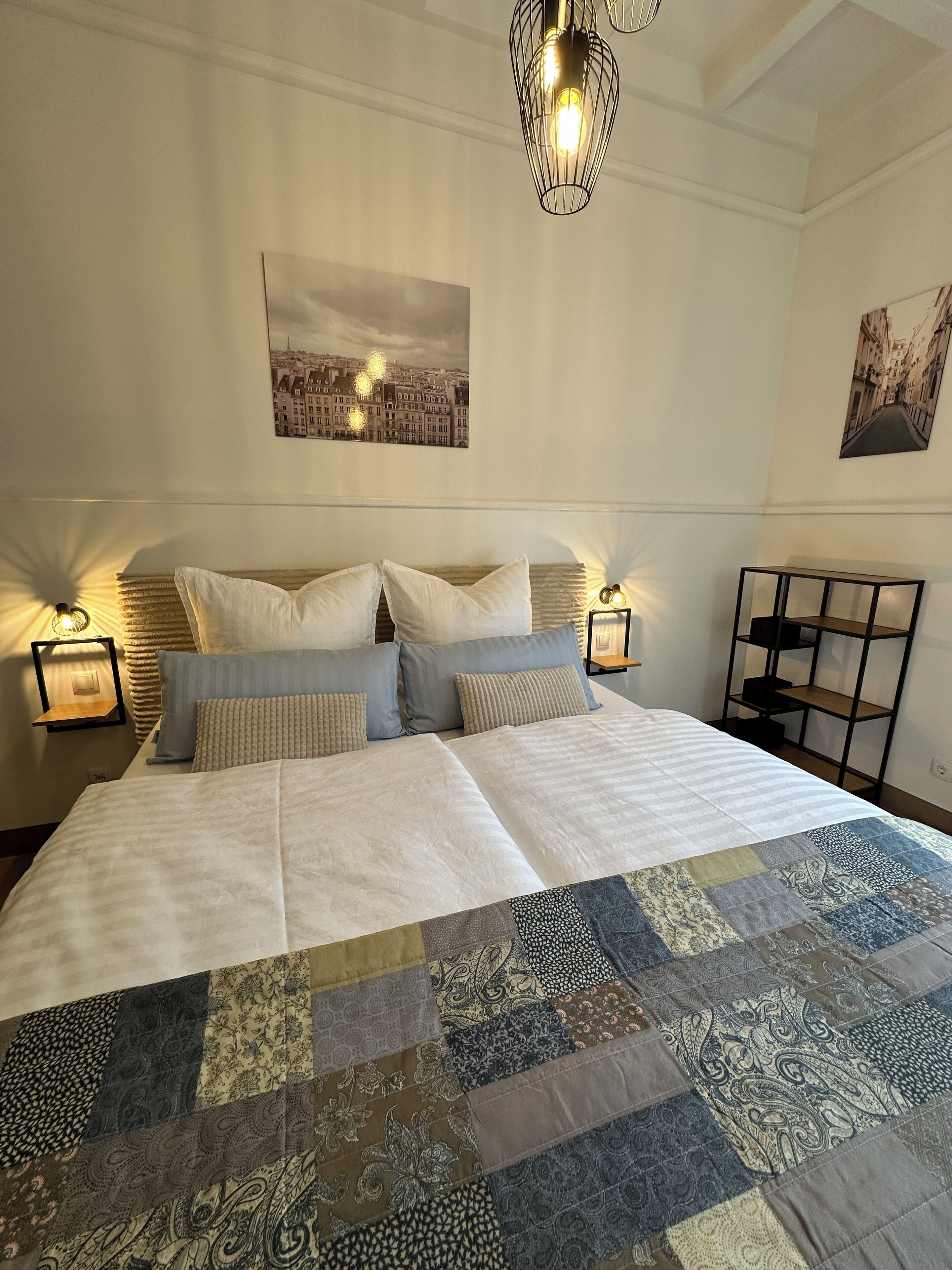 Ein ordentlich bezogenes Bett mit einem Patchwork-Quilt, zwei Nachttischlampen und einem Stadtbild darüber.
