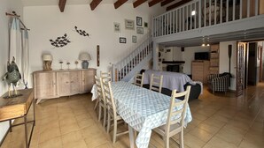 Une salle à manger spacieuse avec un plafond haut et des poutres apparentes, un escalier menant à un niveau supérieur et une vue sur la cuisine.