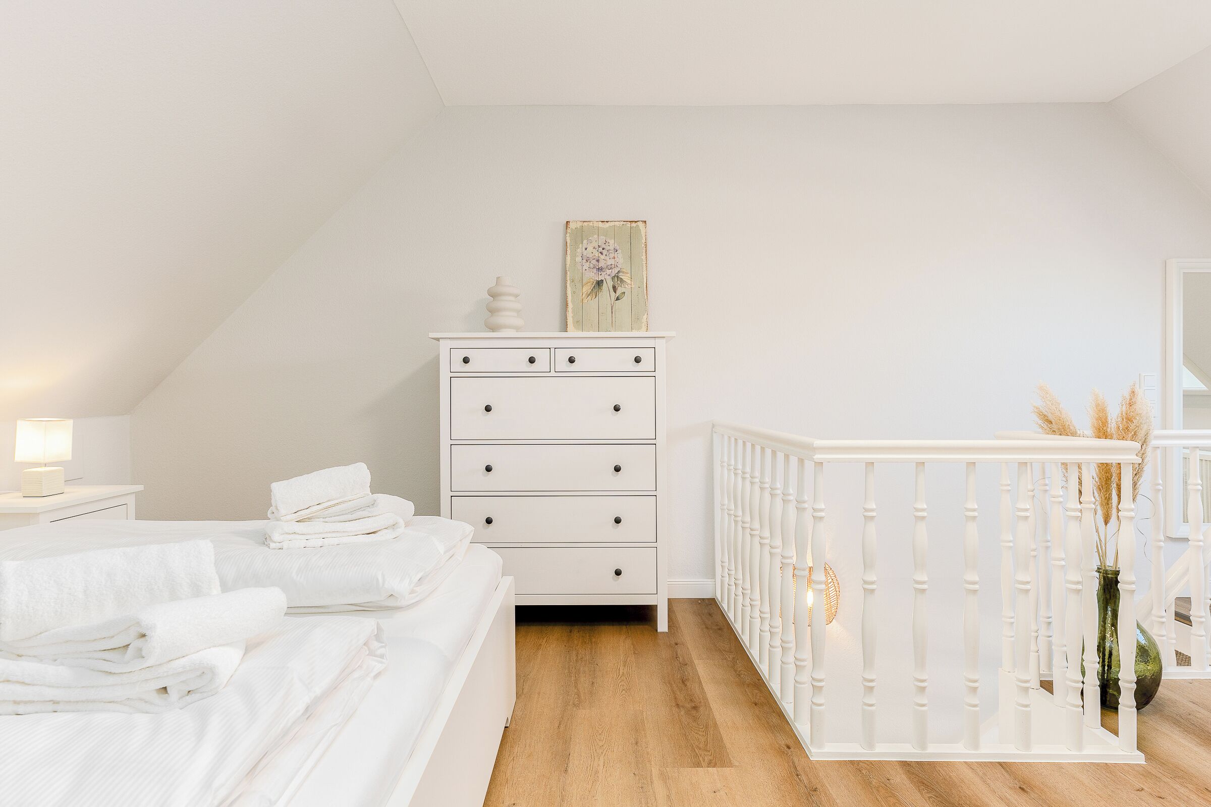 A bedroom with a white dresser, a bed with neatly folded white towels, a white crib, and a wooden floor.