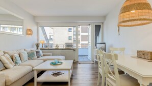 A modern living room with a sofa, coffee table, and dining table. There is a large window providing a view of the outside. - Jonathan - House Winora - Westerland (Westerland)