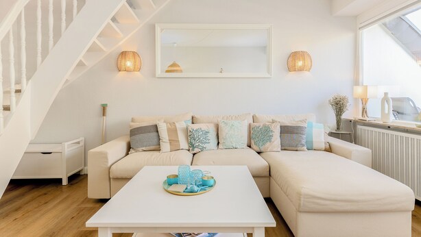 A modern living room with a white staircase, a beige sofa with patterned cushions, a white coffee table with decorative items, and a large window providing natural light. - Jonathan - House Winora - Westerland (Westerland)